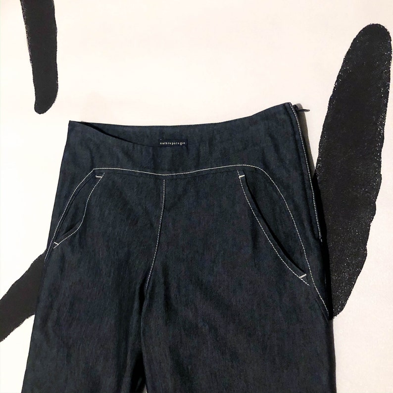 May include: Dark blue denim pants with white stitching detail. The pants feature front pockets and a zipper closure. The brand name "anthropologie" is visible on the waistband. The pants are displayed against a white background with black abstract shapes.