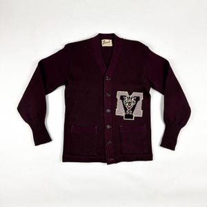 Könnte beinhalten: Ein dunkelroter Strickpullover mit einem großen gestickten Aufnäher auf der Vorderseite. Der Aufnäher zeigt eine weiße Wildkatze mit einem schwarzen 'M' und der Zahl '52' in Schwarz.
