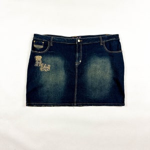 May include: Dark wash denim skirt with a faded design. Features a button closure, front pockets, and decorative gold-colored embellishments including a crown and the letters "RWA".
