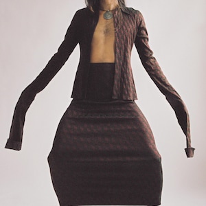 May include: A burgundy two-piece outfit with a geometric pattern. The ensemble includes a long-sleeved jacket and a matching skirt. The model is wearing white boots and a silver pendant necklace.