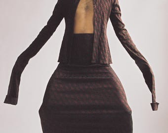 May include: A burgundy two-piece outfit with a geometric pattern. The ensemble includes a long-sleeved jacket and a matching skirt. The model is wearing white boots and a silver pendant necklace.