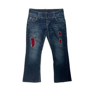 May include: Blue denim jeans with distressed patches featuring red fabric underneath. The jeans have a button and zipper closure, pockets, and a slight flare at the bottom. The jeans have a faded wash and visible stitching.