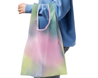 May include: A reusable shopping bag with a colorful tie-dye design. The bag features a gradient of pink, green, and yellow hues. The bag's trim is a soft pink color. The bag is held by a person wearing a blue hoodie.
