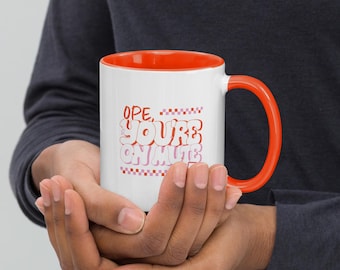 May include: White ceramic mug with an orange interior and handle. The mug features the text "Ope, You're on Mute" in a retro font with pink and red accents. The mug is held in both hands.