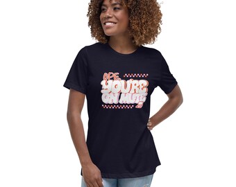 May include: Navy blue t-shirt with the text "Ope, You're on Mute" in a white, orange, and red bubble font. The text is framed by a checkered pattern. The shirt is worn by a person.