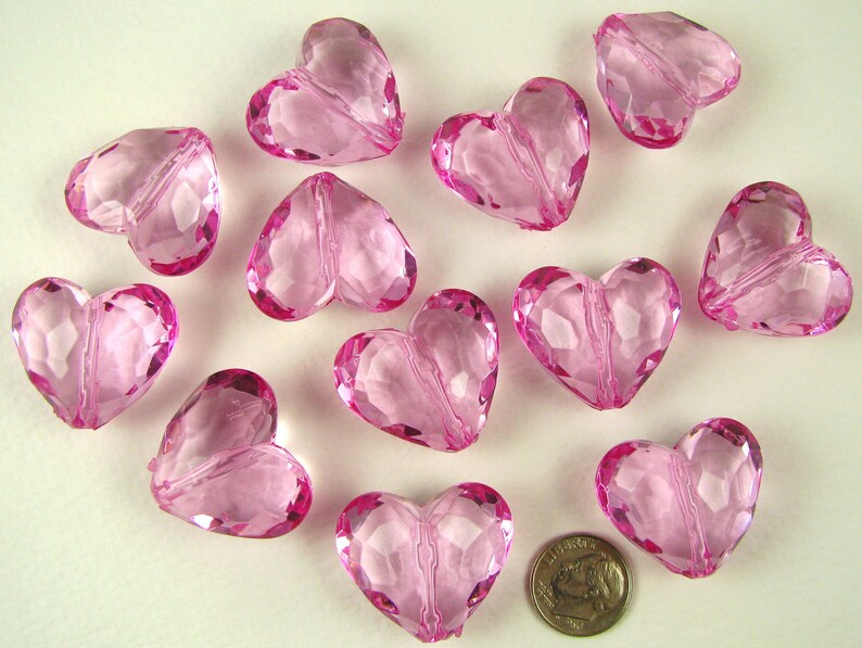 May include: A collection of twelve pink heart-shaped beads. The beads are made of clear acrylic and have a faceted design.