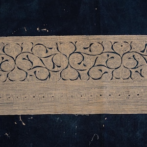 Könnte beinhalten: Cremefarbener Stoff mit einem detaillierten, sich wiederholenden Muster aus wirbelnden Ranken und floralen Elementen. Das Design wird von einem geometrischen Muster begrenzt. Der Stoff ist vor einem dunkelblauen Hintergrund platziert.