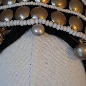 May include: Close-up of a decorative headpiece featuring silver-toned metal discs, small bells, and white beaded strands. The headpiece is set against a black fabric base and a white fabric background.