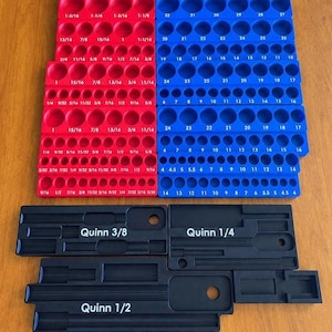 May include: A red and blue plastic socket organiser tray set with printed fractional and metric size labels. Below the trays are three black plastic organisers labelled Quinn 3/8, Quinn 1/4, and Quinn 1/2, featuring recessed slots for tools.