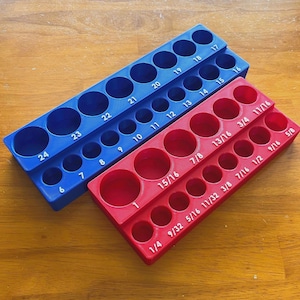 May include: A blue and a red tiered drill bit organiser tray. The blue tray features numerical markings from 6 to 24. The red tray features fractional inch markings ranging from 1/4 to 1. Each tray has circular slots for holding drill bits in an organised manner.