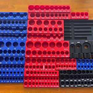 Puede incluir: Una colección de organizadores de herramientas multicolores en azul, rojo y negro. Estos bloques rectangulares presentan numerosos recortes circulares de varios tamaños, cada uno etiquetado con medidas imperiales como 1/2, 3/4 y 1-1/4 pulgadas.