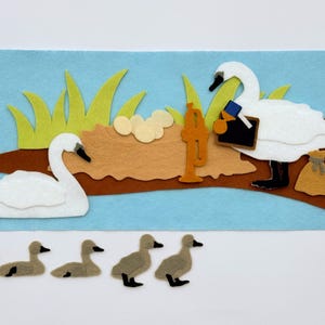 May include: A felt craft depicting a scene with swans and ducklings. Two white swans are on a brown branch with a nest, eggs, and tall green grass. Five gray ducklings are below. A blue background completes the scene.