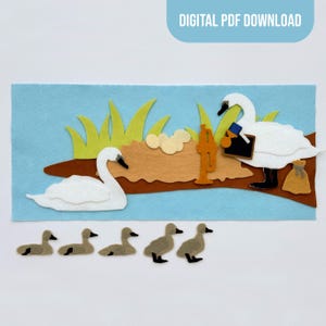 May include: A felt craft set featuring two white swans, five small brown ducklings, green grass, a brown nest with eggs, a gold trumpet, a black chalkboard with a blue frame, and a small tan sack, all set against a light blue background.
