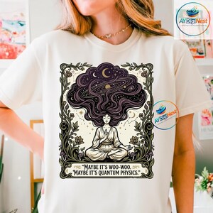 Può includere: T-shirt color crema con grafica dettagliata. Il design presenta una figura che medita con i capelli che si trasformano in una scena cosmica di stelle, pianeti e una luna crescente. Il testo recita: "MAYBE IT'S WOO-WOO. MAYBE IT'S QUANTUM PHYSICS."