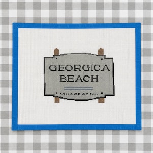 May include: A needlepoint canvas featuring a sign that reads "GEORGICA BEACH" in black text. The sign is gray with brown posts and a blue border. The text "VILLAGE OF E.H." is below the beach name.