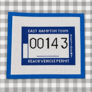 May include: A blue and white needlepoint Beach Vehicle Permit from East Hampton Town. The permit features the number "00143" in black, with the word "Resident" on the right side. The permit is framed with a blue border.
