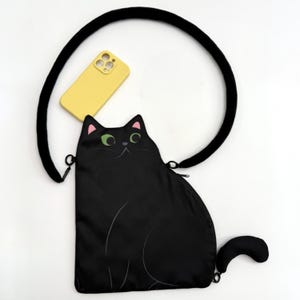 May include: Black cat-shaped crossbody bag with a black strap and a yellow phone case. The bag features a cat face with green eyes and pink ear accents. The bag's tail is curved. The strap is thick and black.