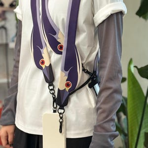 May include: A long, flat phone strap featuring a repeating graphic of a bird head with a dark purple neck, a cream-coloured face, and a red eye. The strap is attached to a plain white smartphone case via black metal clips and a small black chain link.