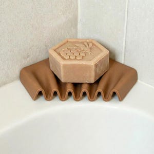 May include: A brown soap dish with a wavy design holds a hexagonal bar of soap. The soap is light brown and features a bee and honeycomb design. The soap dish is placed in the corner of a white bathtub.