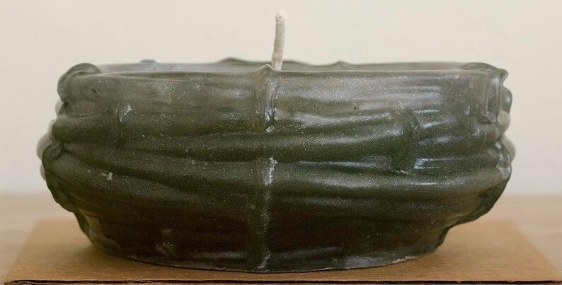 May include: A dark green, textured candle in a bowl shape. The candle has a thick, woven-like design around the sides and a single, unlit wick in the center. It sits on a brown cardboard surface.