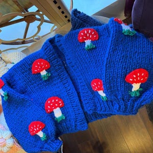 May include: A bright blue knitted cardigan featuring red mushroom decorations with white stems and green bases. The open-front sweater has long sleeves and a chunky knit, offering a playful and unique style.