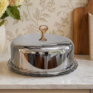 May include: A shiny, silver cake stand with a domed lid and a decorative handle. The stand has a scalloped edge and sits on a marble countertop. The handle is gold-colored and ornate. The background includes a floral wallpaper and a wooden cutting board.