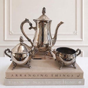 May include: A silver-toned tea set consisting of a tall teapot with a lid, a lidded sugar bowl, and a creamer. Each piece features ornate, curved handles and decorative feet. The set is arranged on two stacked books with visible text on the spines.