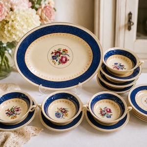 May include: A ceramic dinnerware set featuring an oval serving platter and several two-handled soup bowls with matching saucers. Each piece has a cream-colored center, a deep blue border, and a central floral bouquet with gold trim accents.