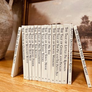 May include: A collection of vintage children's books with white spines and gold lettering. Titles include "The Tale of Peter Rabbit" series. The books are arranged on a wooden surface, with a framed picture in the background.