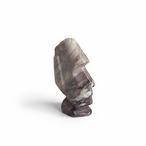 Puede incluir: Una escultura de piedra tallada de una cabeza Moái, vista de perfil. La escultura es principalmente de tonos morados y blancos, con textura y detalles visibles. La cabeza está sobre un fondo blanco.
