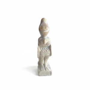 Può includere: Una statuina in pietra scolpita di una figura simile a un uccello. La scultura ha una testa arrotondata con una cresta, un petto dettagliato e una vita decorata. La figura è su una base rettangolare ed è di colore grigio chiaro.