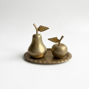 Peut inclure: Un ensemble de salière et poivrière en laiton. L'ensemble comprend un shaker en forme de poire et un shaker en forme de pomme, chacun avec un détail de feuille sur le dessus. Ils reposent sur un plateau rond en laiton avec un bord décoratif.