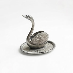 May include: A silver-toned swan figurine with detailed feather and wing designs, perched on a matching oval tray. The swan's neck is gracefully arched, and the tray features an ornate, embossed pattern. A decorative piece.