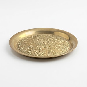 May include: A round, gold-colored brass tray with an ornate, embossed floral design. The tray has a slightly raised edge and a polished finish, suitable for decorative use or serving.