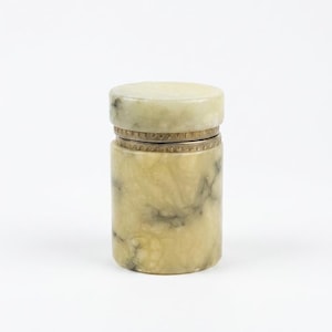 May include: A cylindrical container with a lid. The container is made of a pale, marbled stone with gray and brown veining. The lid and a band around the middle have a decorative, textured design. The container is on a white background.
