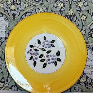 May include: A round, yellow ceramic plate with a white center featuring a botanical design. The design includes a branch with purple berries and green leaves. The plate has a diameter of approximately 10 inches.