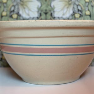 May include: Cream-colored ceramic mixing bowl with a wide rim and three horizontal stripes. The stripes are a combination of light blue and pink, adding a decorative touch to the kitchenware. The bowl is a classic design.
