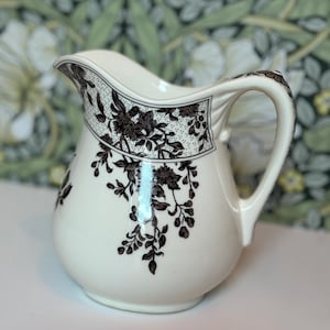 May include: Cream-colored ceramic pitcher with a black floral design. The pitcher features a curved spout and a handle. The floral pattern includes flowers and leaves. The pitcher is likely used for serving beverages.