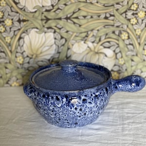 May include: A blue ceramic soup bowl with a lid and handle. The bowl has a textured, speckled design with white accents. The lid has a small knob. The bowl is on a white surface.