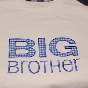 May include: White t-shirt with the words "BIG BROTHER" embroidered in blue. The letters "BIG" are filled with a blue and white gingham pattern, creating a contrasting design element. The text is centered on the shirt.