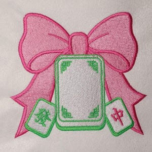 Puede incluir: Un diseño bordado que presenta un gran lazo rosa centrado detrás de una ficha de mahjong rectangular con cara blanca en blanco y borde verde. Dos fichas de mahjong más pequeñas se colocan en la parte inferior, una con un carácter verde y otra con uno rojo.