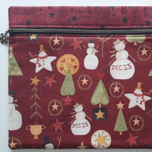 Pode incluir: Uma bolsa retangular de tecido com zíper em vermelho escuro com um padrão repetitivo de bonecos de neve, árvores de Natal verdes e estrelas. Alguns bonecos exibem o texto DEC 25. A borda superior inclui PEACE ON EARTH GOOD WILL TO ALL.