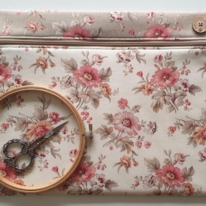 May include: A beige floral pattern zip pouch with a wooden button closure. A wooden embroidery hoop holds a pair of silver scissors. The pouch and hoop feature a cream background with pink and brown floral designs.