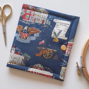 May include: A navy blue fabric needlework case with a patriotic print featuring buildings, an eagle, and a scroll. The case is secured with a gold snap closure. Gold scissors and a wooden embroidery hoop are visible.