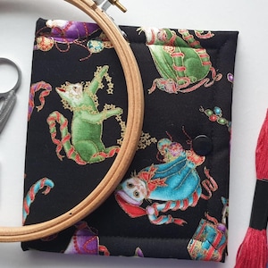 May include: A black fabric needlework organizer featuring a colorful cat print, a wooden embroidery hoop, silver metal stork scissors, and two skeins of embroidery floss in red and blue. The organizer has a black snap button closure.