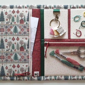 Pode incluir: Um organizador de artesanato em tecido com estampa natalina de casas vermelhas, árvores verdes e ornamentos. Inclui um bolso com zíper, um alfineteiro de feltro com linha de bordar, uma tesoura pequena e duas meadas.
