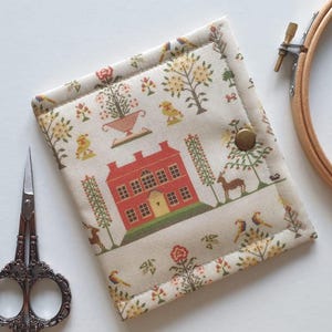 May include: A fabric needlework case with a red house and floral pattern. The case is closed with a brass snap. A pair of ornate scissors and an embroidery hoop are next to the case. The case is beige with a colorful print.