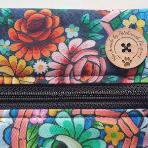 May include: Close-up of a floral patterned pouch with a black zipper. The fabric features a dark blue background with orange, pink, yellow, and white flowers. A wooden button with the text "handmade by patchwork pawprint" is attached.