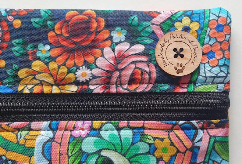 May include: Close-up of a floral patterned pouch with a black zipper. The fabric features a dark blue background with orange, pink, yellow, and white flowers. A wooden button with the text "handmade by patchwork pawprint" is attached.