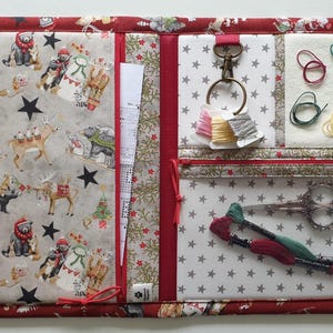 May include: A fabric needlework organizer featuring a red border with a holiday animal print. The interior includes a pocket with a winter forest pattern, a white star-print section with a metal ring, embroidery floss, scissors, and thread.